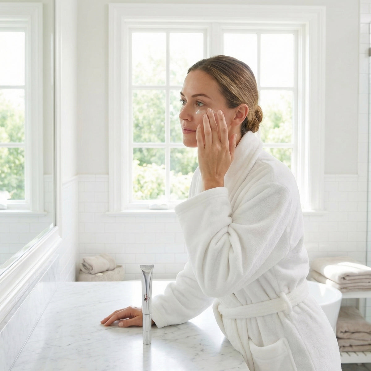 Woman in a white robe applying cream to her face in a bright, white room.